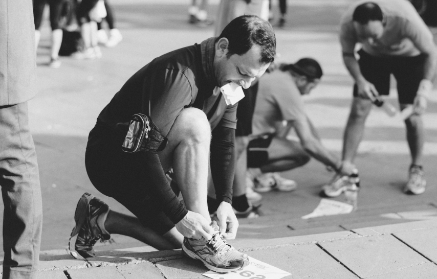 A runner on bended knee stops to tie his running shoes with a document or bill in his mouth